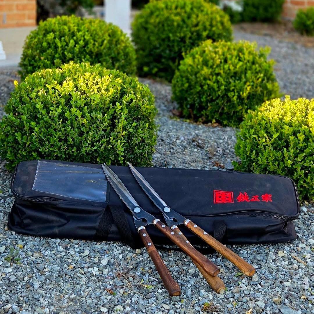 Yoshioka garden shear and tool bag placed in a garden with two hedge shears leaning against it in front of a round trimmed Buxus ball